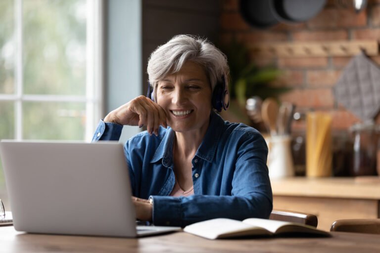 A middle-aged woman wearing a headset smiles at her computer as if enjoying an online class.