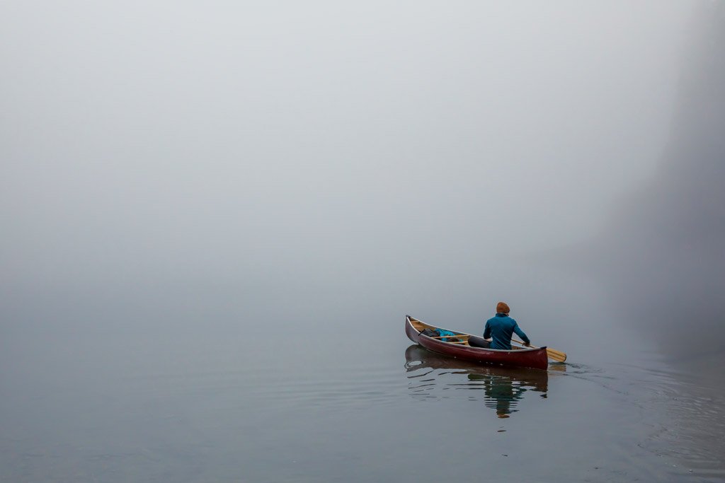 One person in a red canoe paddles away from the camera on a very foggy day. No shoreline is visible.